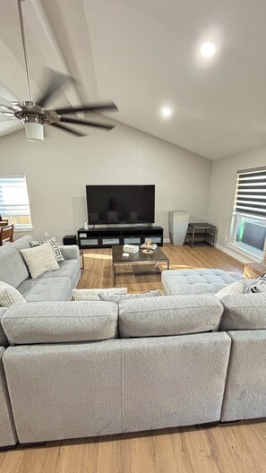A modern living room with a sectional sofa, a coffee table, a flat-screen TV, and a ceiling fan. - Cozy Katy Home near Katy Mills, Restaurants, and Family Attractions (Katy)