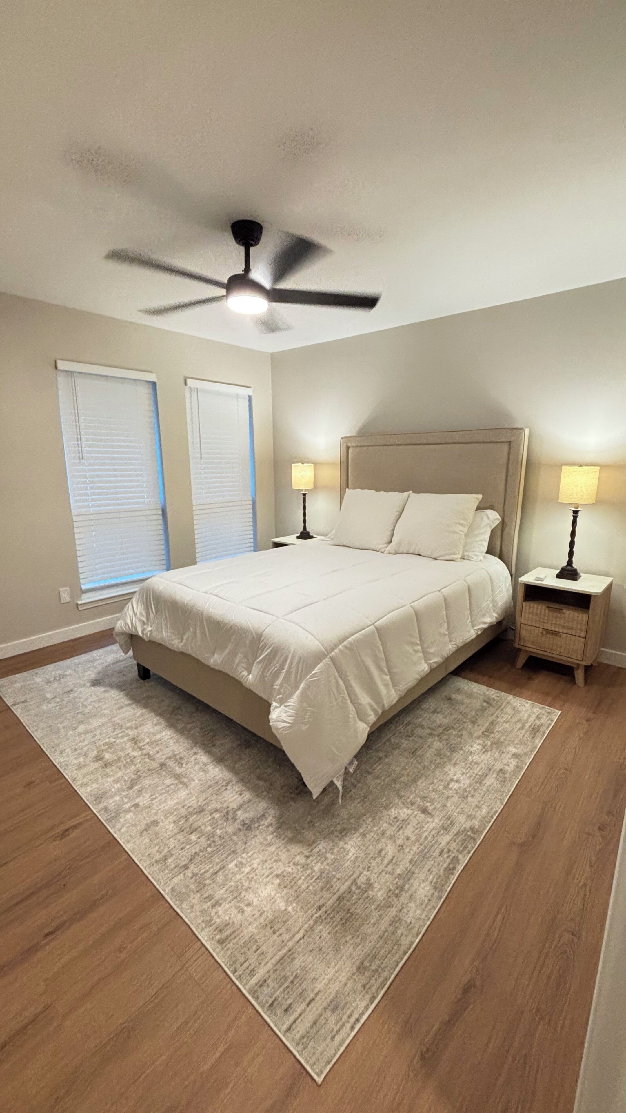 A bedroom with a bed, a ceiling fan, two bedside lamps, a nightstand, and a window with blinds.