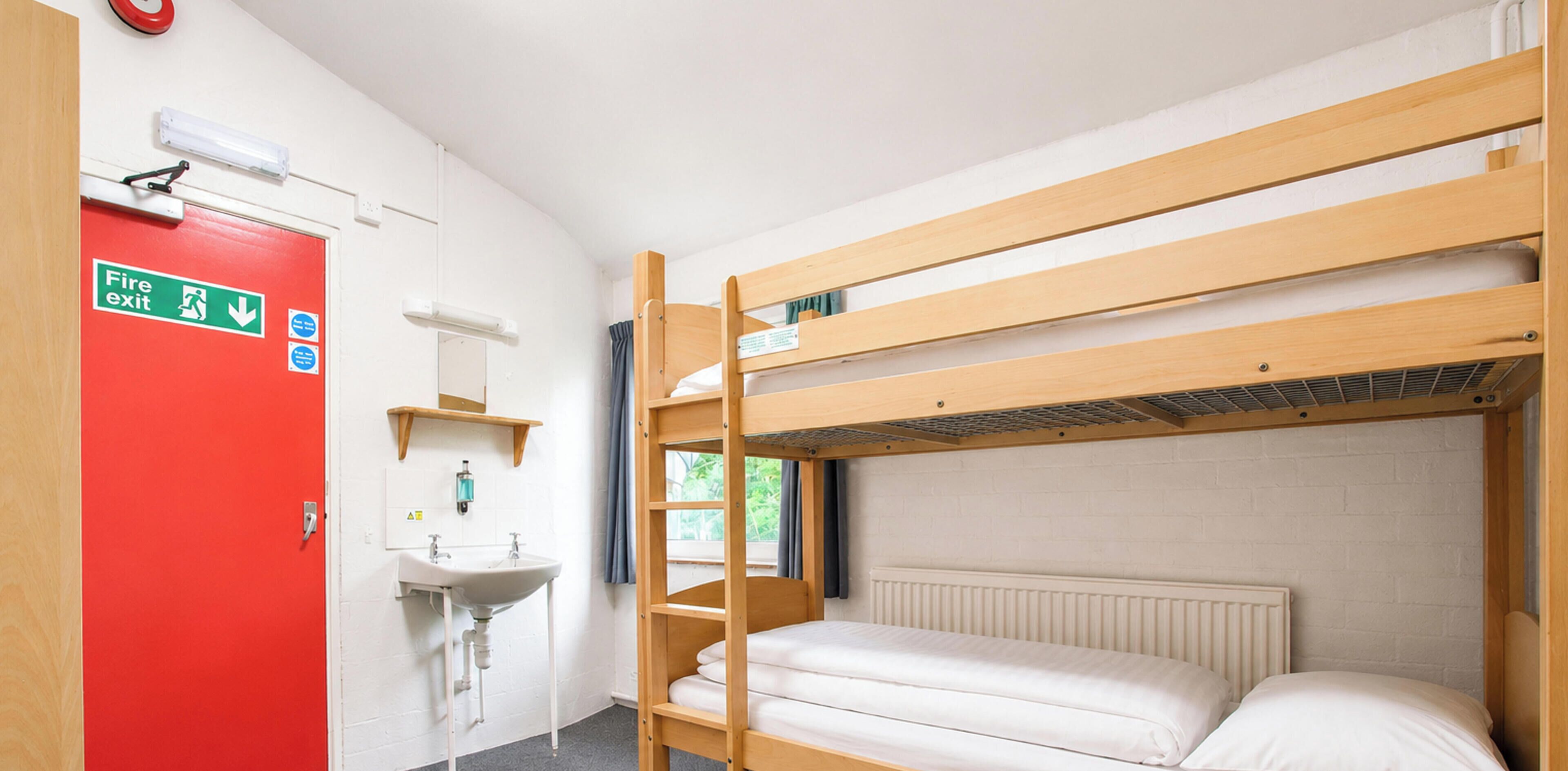 A bunk bed room with a red door marked 'Fire exit,' a white sink, and a window with curtains.