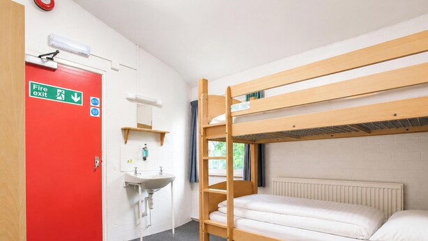 A bunk bed room with a red door marked 'Fire exit,' a white sink, and a window with curtains.