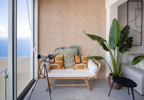 En balkon med et teleskop, en fletningssofa med hynder, en stor plantepotte og udsigt til havet.