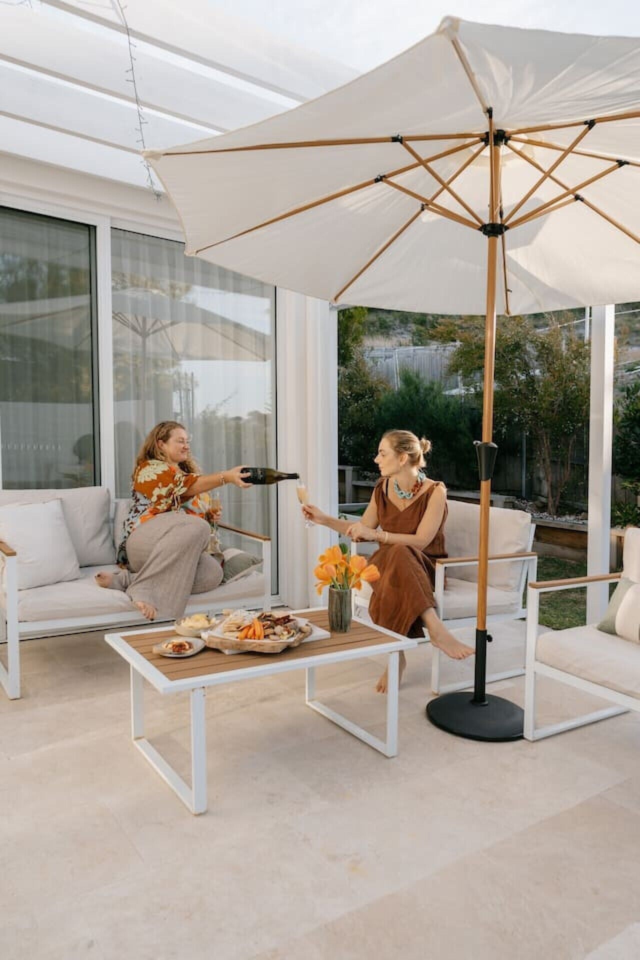 An outdoor seating area with a white umbrella, a table with food, and two people toasting.