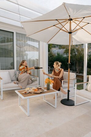 An outdoor seating area with a white umbrella, a table with food, and two people toasting.