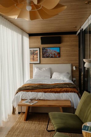 A modern bedroom with a wooden headboard, a bed with white bedding, a green armchair, and a large window with sheer curtains. - Casa Collection Bicheno ~ The Bambino (Bicheno)