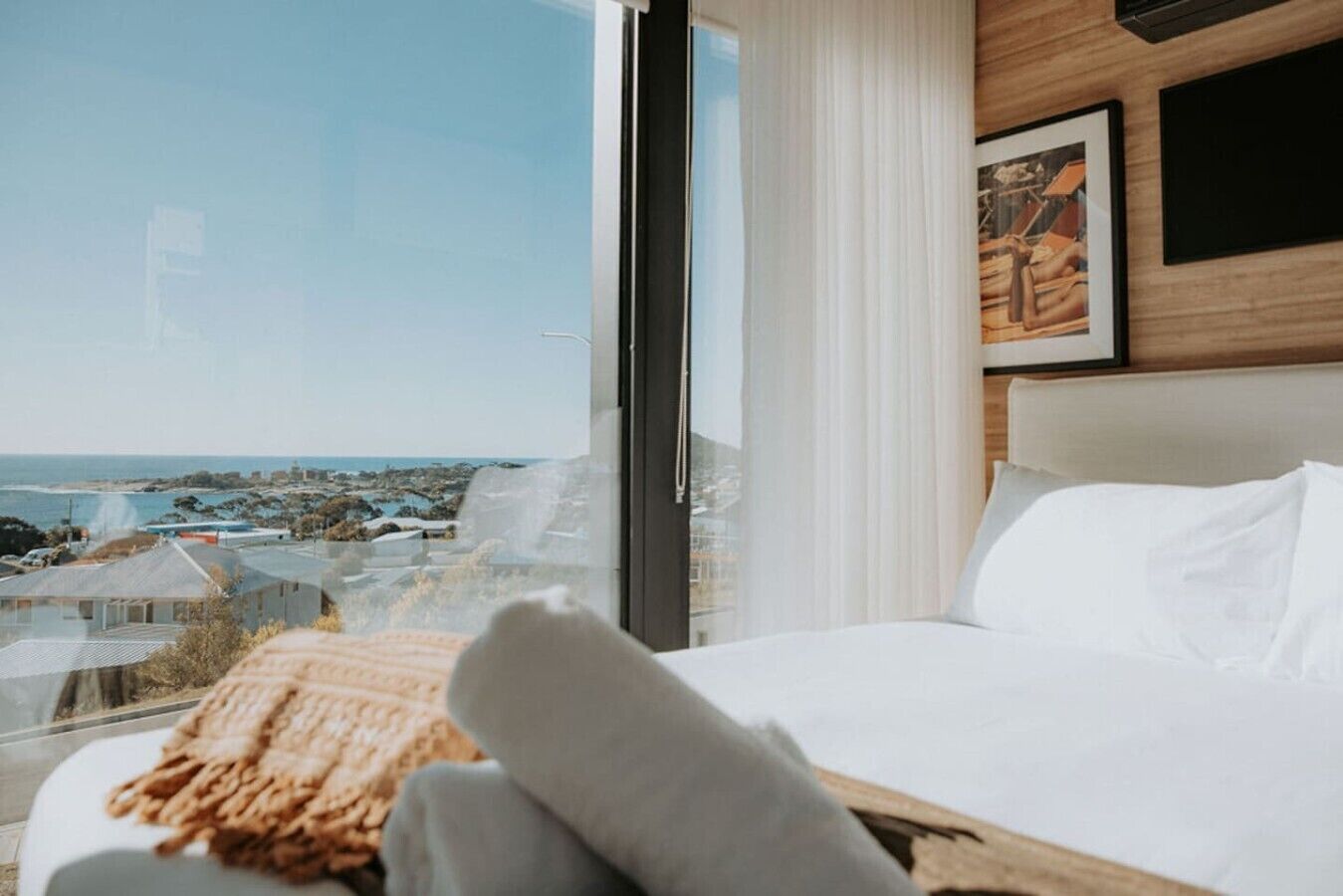 A hotel room with a large window, a bed with white linens, a wooden headboard, and a framed picture on the wall.