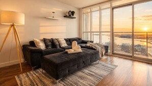 A modern living room with a dark sofa, a floor lamp, and a large window offering a city view. - Waterfront Grand Escape (Toronto)
