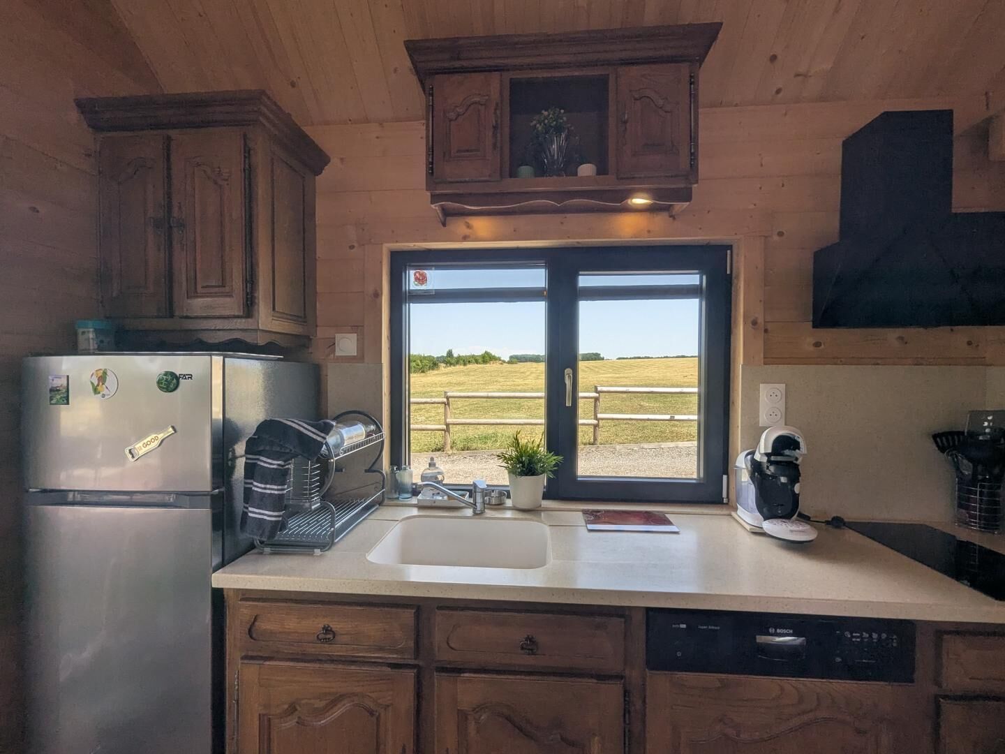 Cocina con armarios de madera, un refrigerador de acero inoxidable, un lavavajillas Bosch y una ventana con vistas a un campo.
