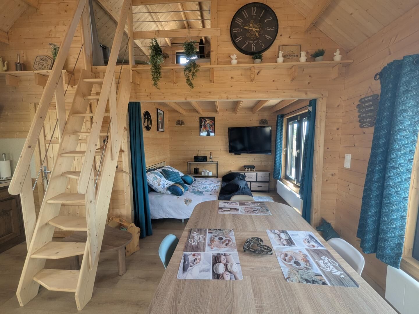 Ein Holzblockhaus-Interieur mit Treppe, einem Bett, einem Esstisch mit Platzsets, einem Fernseher und einer Wanduhr.