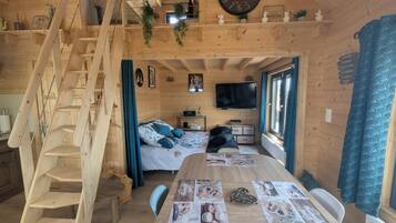 Ein Holzblockhaus-Interieur mit Treppe, einem Bett, einem Esstisch mit Platzsets, einem Fernseher und einer Wanduhr.