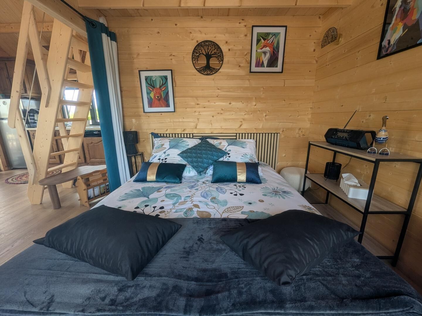 A wooden cabin interior with a bed, a ladder, a shelf with a radio, and framed artwork on the walls.