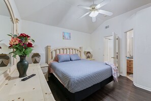Een slaapkamer met een bed, nachtkastje, vaas met bloemen, afstandsbediening, plafondventilator en een deur naar een andere kamer.