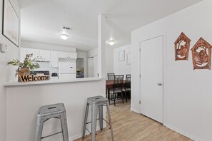 Moderni keittiö, jossa on valkoiset kaapit, sisäänrakennettu uuni sekä ruokailutila, jossa on puinen pöytä ja tuolit.