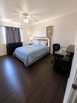 A bedroom with a bed, a desk, a chair, a ceiling fan, and a window with curtains.