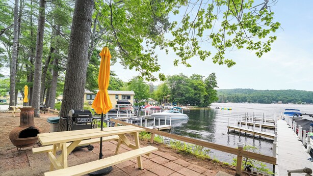 A lakeside picnic area with wooden benches, a grill, and umbrellas. - Cabin 42 at the Historic Sandy Point Resort W/ Boat Slip and Onsite Boat Ramp (Alton)