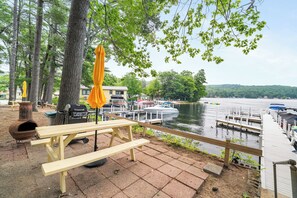 A lakeside picnic area with wooden benches, a grill, and umbrellas. - Cabin 42 at the Historic Sandy Point Resort W/ Boat Slip and Onsite Boat Ramp (Alton)