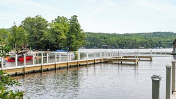 A calm lake with a wooden dock extending into the water, surrounded by lush greenery and a forested hill in the background.