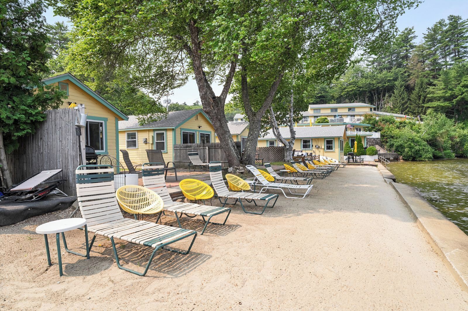 A row of beach chairs and tables along a sandy path leading to a body of water, with trees and buildings in the background.