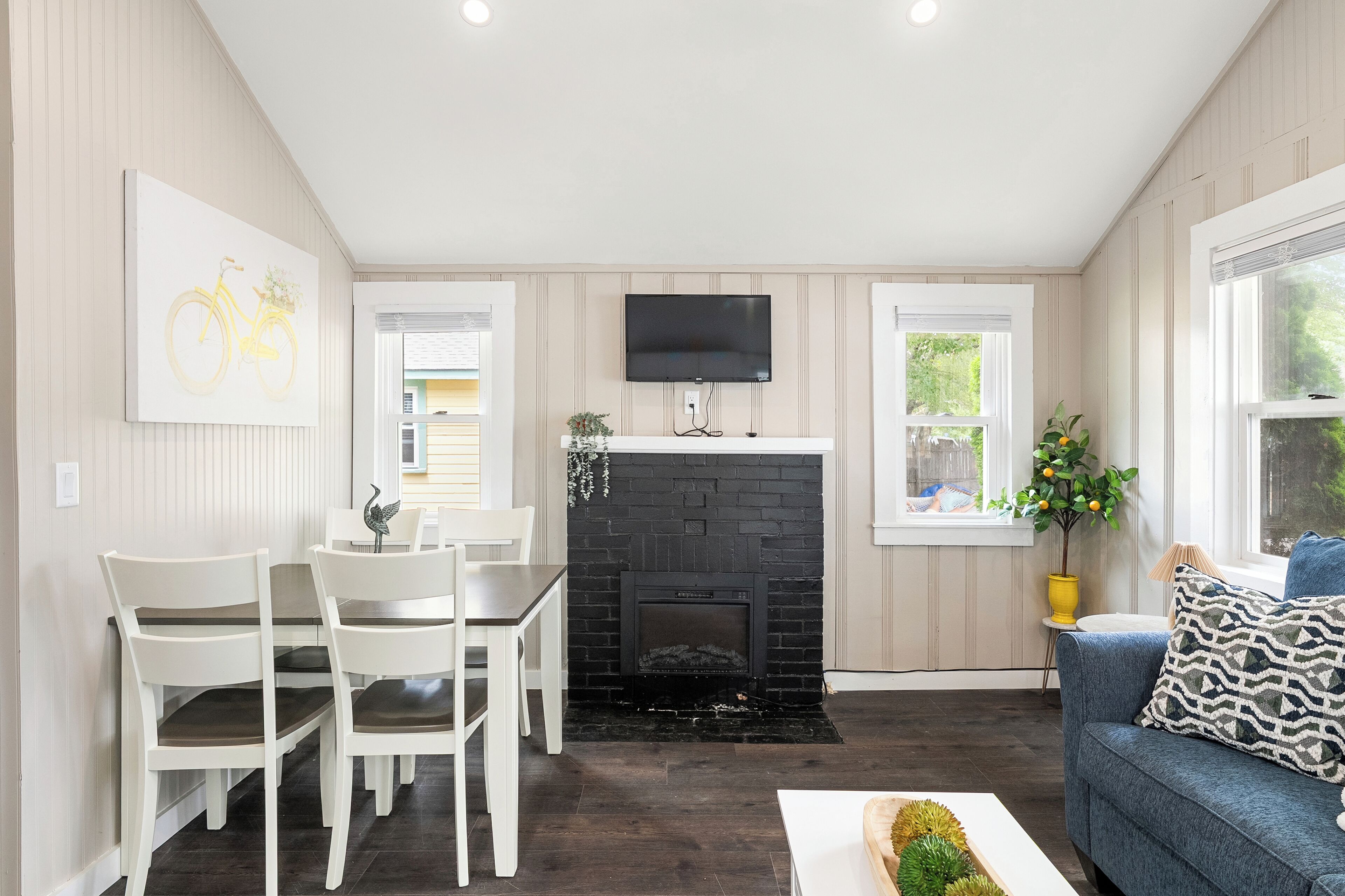 A modern living room with a fireplace, a dining area with white chairs, a flat-screen TV mounted above the fireplace, and a window with a view of greenery.