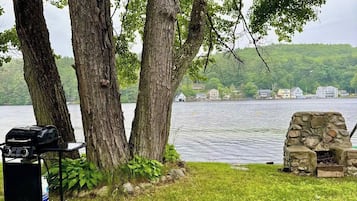A lakeside setting with a grill and stone fireplace, surrounded by trees and a view of buildings across the water.