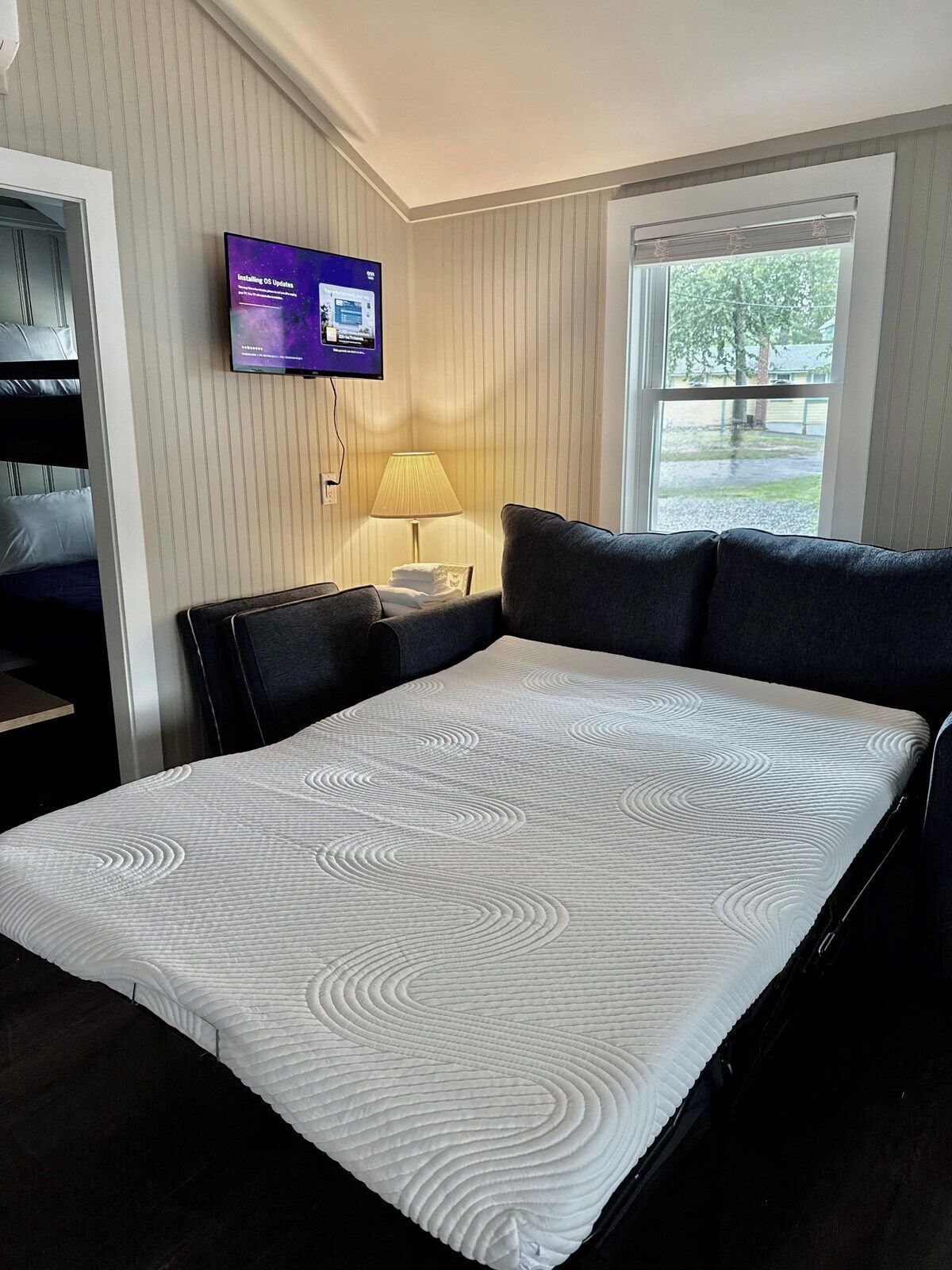 A hotel room with a bed, a television mounted on the wall, a window with a view of trees, and a lamp on a bedside table.