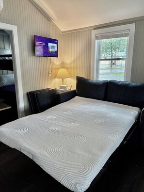 A hotel room with a bed, a television mounted on the wall, a window with a view of trees, and a lamp on a bedside table. - Cabin 59 at Historic Sandy Point Resort W/ Boat Slip and onsite Boat ramp (Alton)