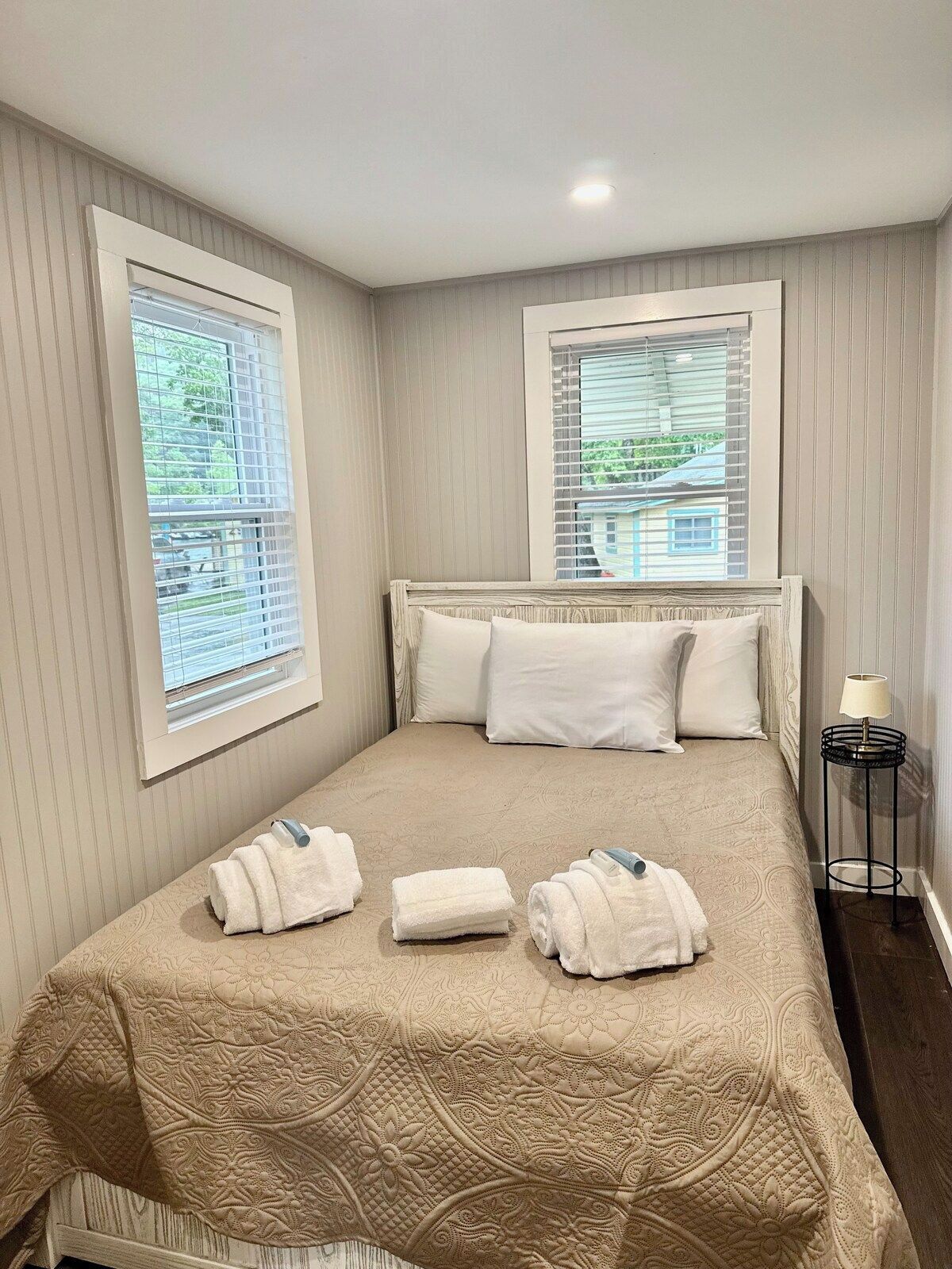 A neatly made bed with a beige bedspread and pillows, a bedside lamp, and two windows with blinds.