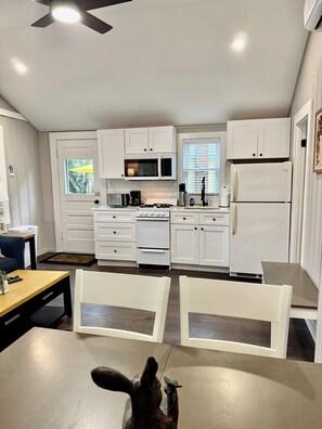 A modern kitchen with white cabinetry, a microwave, a stove, and a refrigerator. There is a dining table with chairs in the foreground. - Cabin 59 at Historic Sandy Point Resort W/ Boat Slip and onsite Boat ramp (Alton)