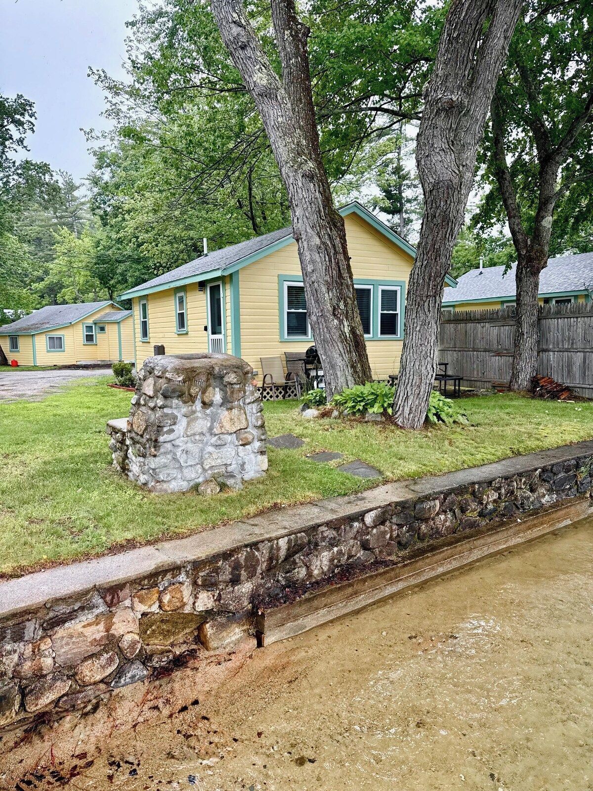 A yellow house with a stone structure in front, surrounded by trees and a wooden fence.
