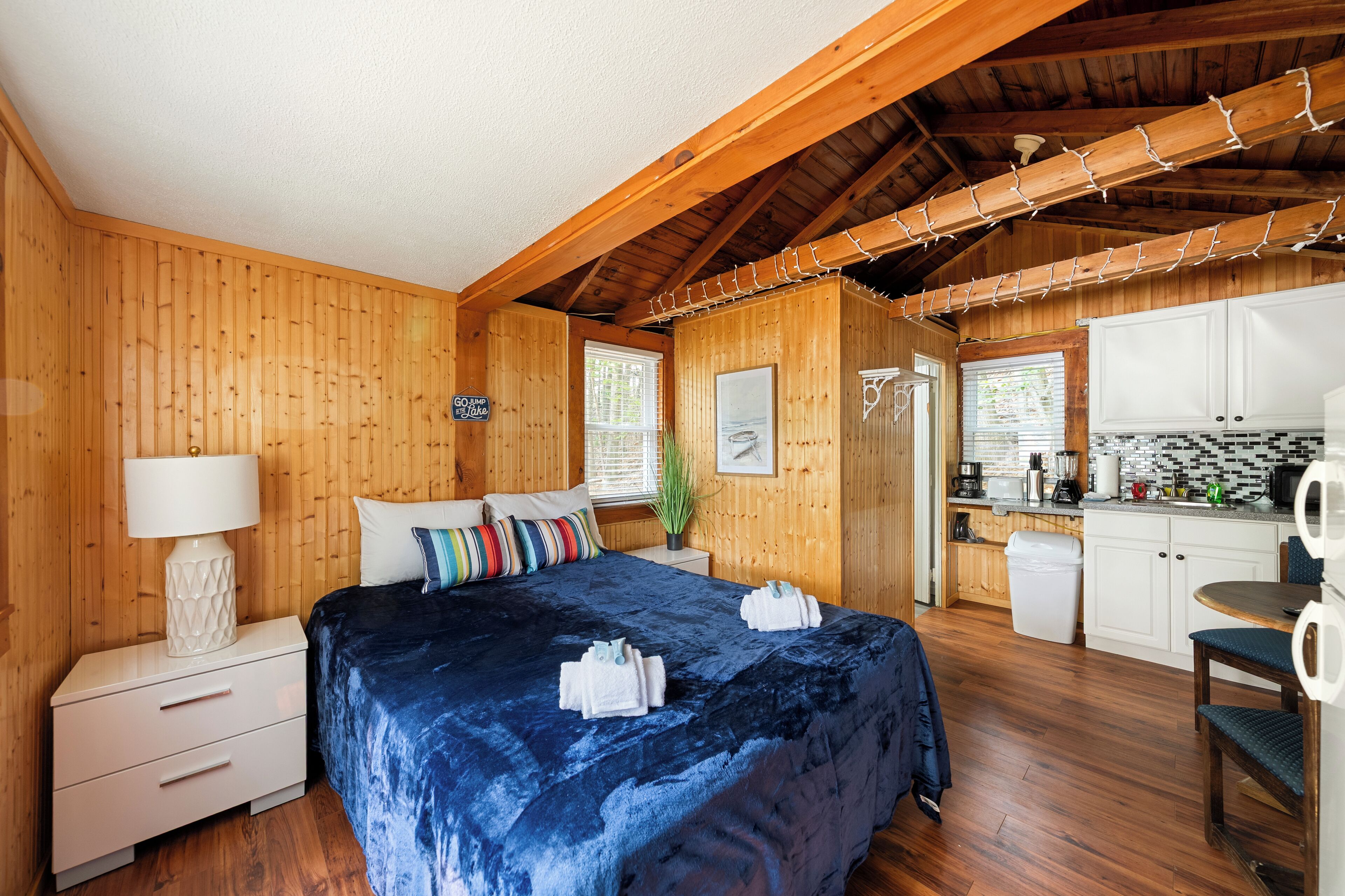 Une cabane en bois comprenant un lit, une table de chevet, une lampe, une fenêtre, un coin cuisine et une table à manger.