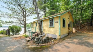 A yellow cabin with a porch, a stone pillar, and a view of a body of water. - Cabin 48 at Historic Sandy Point Resort W/ Boat Slip and Onsite Boat Ramp (Alton)