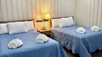 A hotel room with two beds, each with a blue quilted bedspread and white pillows. There is a nightstand with a lamp between the beds.