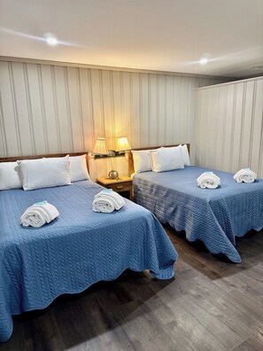 Ein Hotelzimmer mit zwei Betten, beide mit blauen gesteppten Bettdecken und weißen Kissen. Dazwischen befindet sich ein Nachttisch mit einer Lampe.