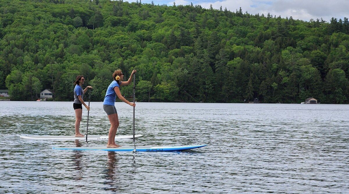 Zwei Personen stehen auf Paddleboards auf einem ruhigen See, im Hintergrund ist ein bewaldeter Hügel zu sehen.