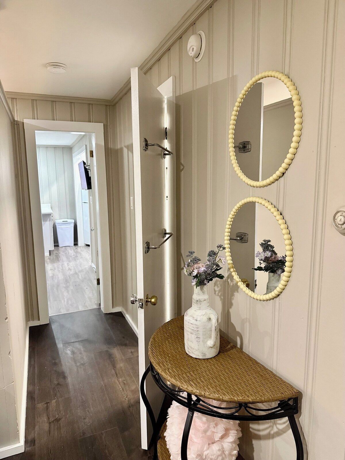 A hallway with a door leading to a bathroom, a mirror with a bead necklace, and a wicker chair with a vase of flowers.