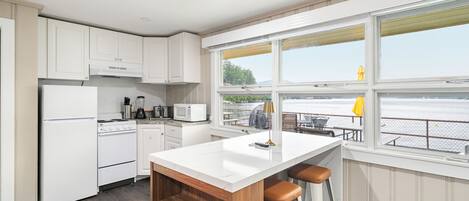 Une cuisine moderne avec des meubles blancs, un îlot central doté de tabourets et une vue sur l’eau grâce à de grandes fenêtres.