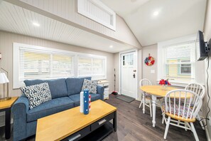 Ein gemütliches Wohnzimmer mit einer Couch, einem Couchtisch und einem Esstisch mit Stühlen. An der Wand befindet sich eine Tür mit einem Kranz, und ein Fenster mit Jalousien.