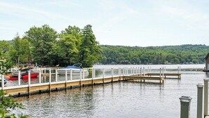 A calm lake with a dock extending into the water, surrounded by trees and a distant hill.
