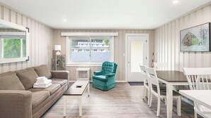 A room with a sofa, a teal armchair, a dining table with chairs, a small side table, and a window with a view of buildings.
