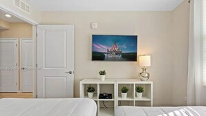 A hotel room with two beds, a TV mounted on the wall, a desk with a lamp, and a painting of a castle. - Storey Lake: Cozy 2BR Near Disney (Kissimmee)