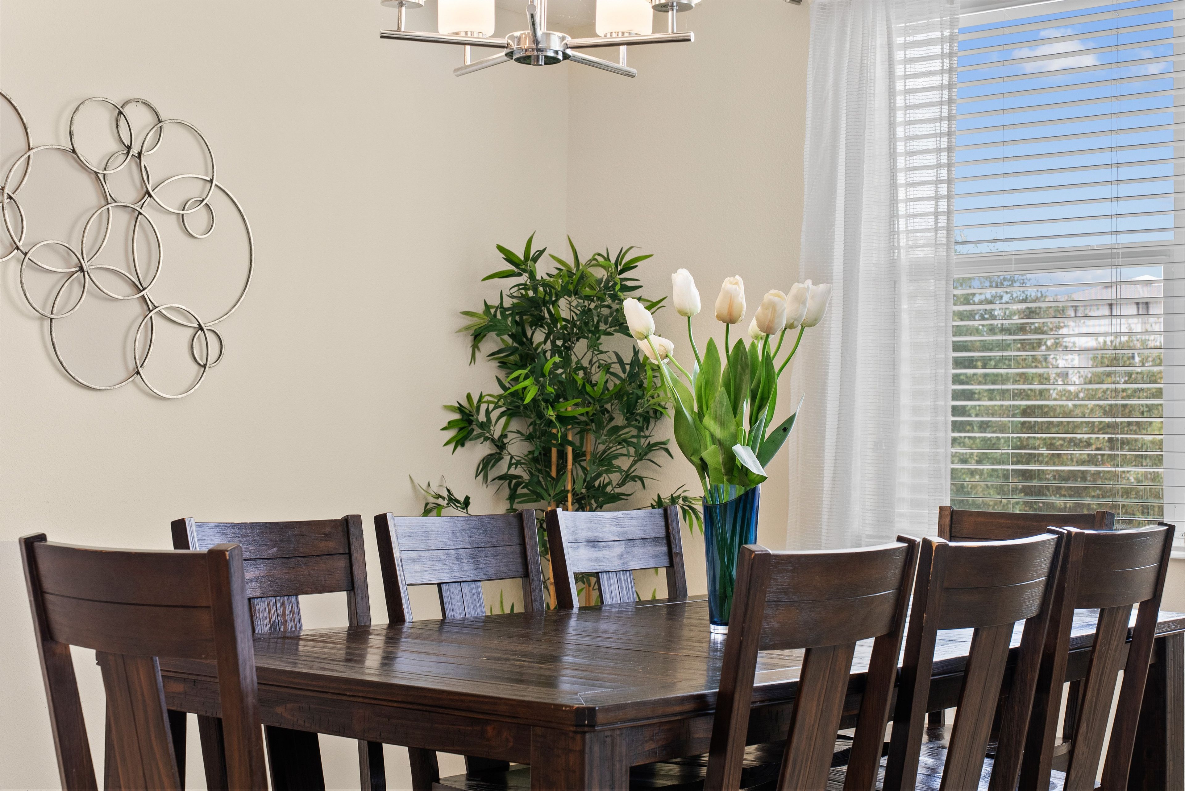 A dining area with a wooden table, chairs, a vase with flowers, a wall-mounted light fixture, and a window with blinds.