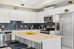 A modern kitchen with white cabinets, a stainless steel refrigerator, a microwave, and a stove. There are two black bar stools at the island. - Storey Lake: Cozy 2BR Near Disney (Kissimmee)