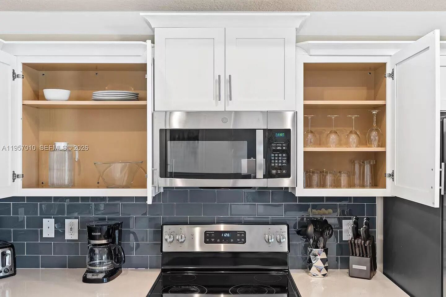 Una cocina con gabinetes blancos, un microondas de acero inoxidable, una estufa negra y una cafetera.