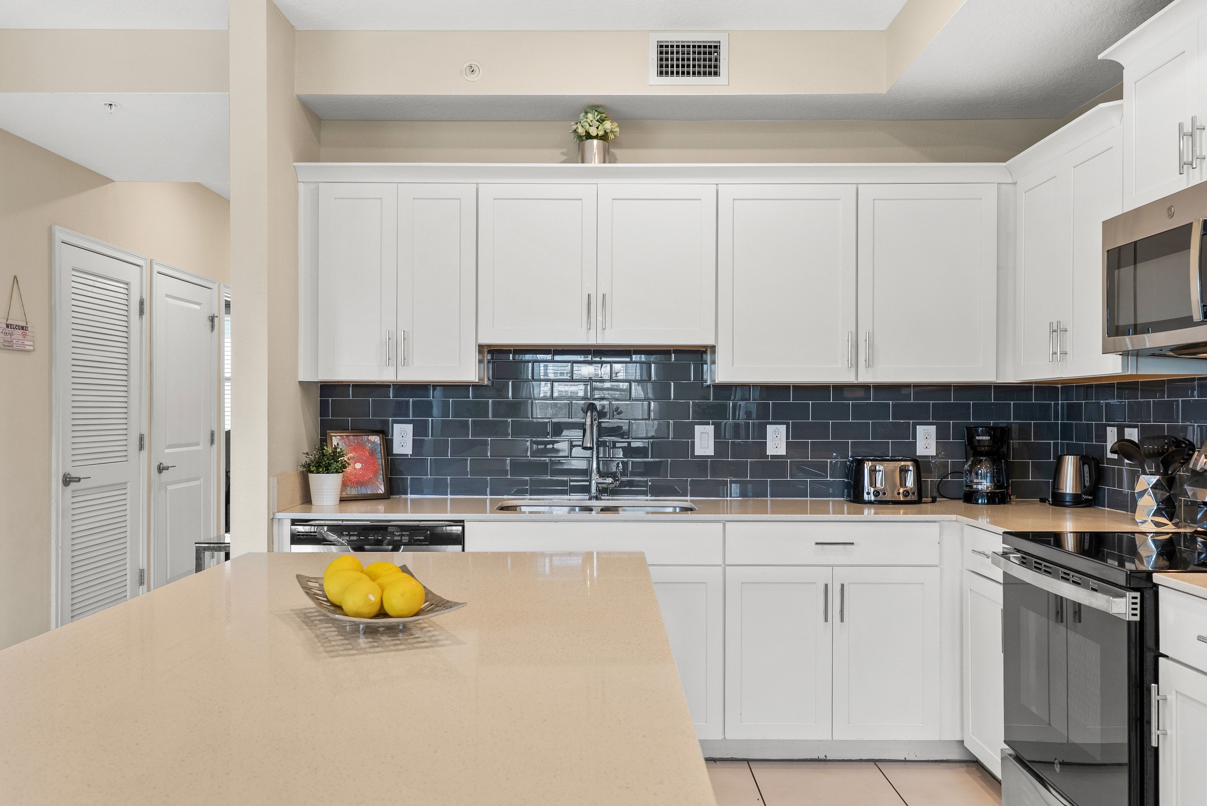 A modern kitchen with white cabinets, a black tiled backsplash, and a central island with a bowl of fruit.