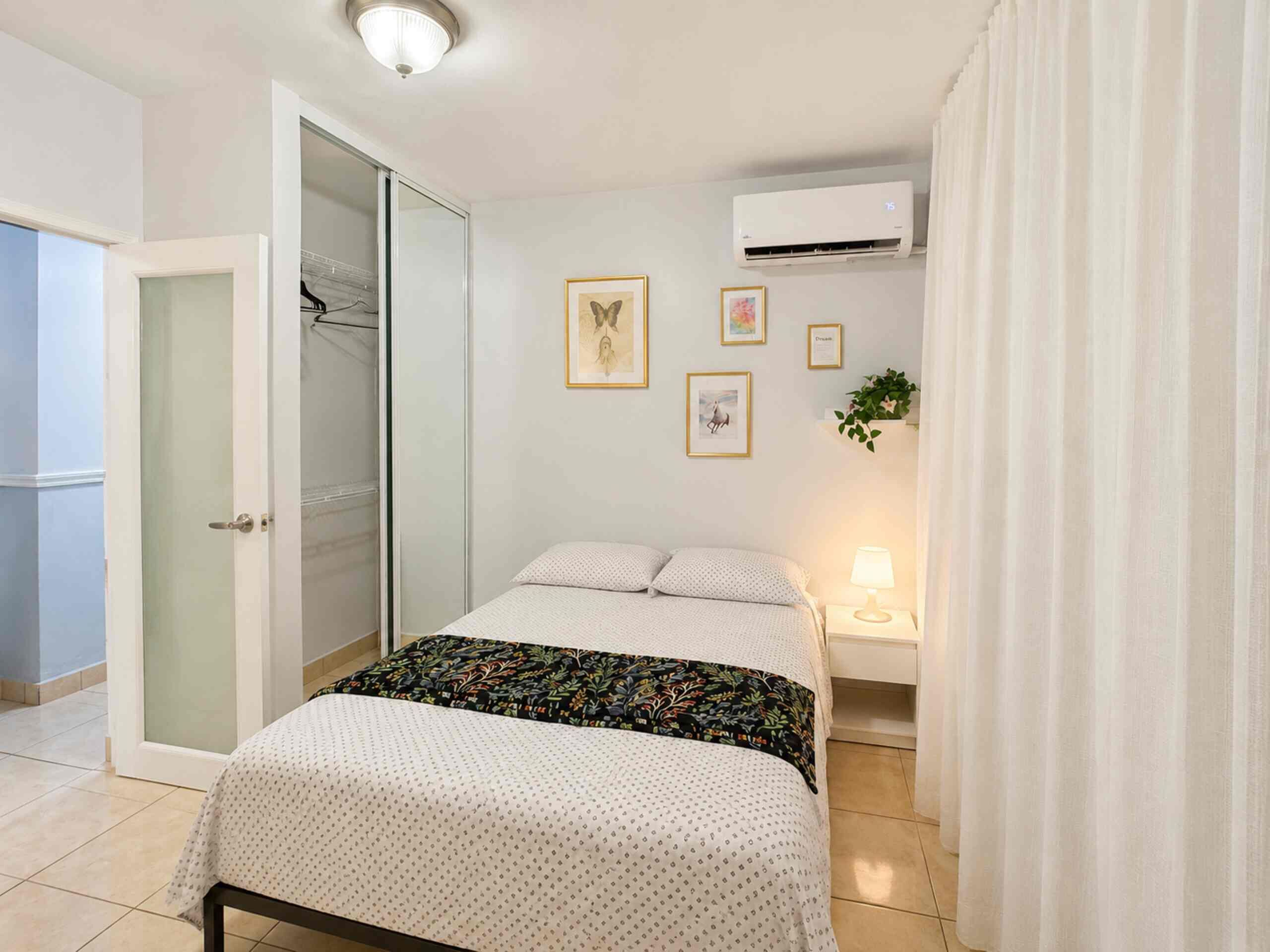 A bedroom with a bed, a nightstand, a lamp, a window with curtains, and an air conditioning unit.
