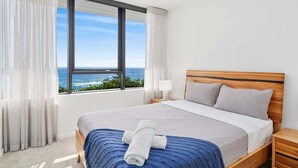 Ett hotellrum med ett stort fönster, en säng med ett blått överkast, ett träfärgat sänggavel och ett nattduksbord med en lampa.