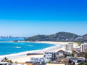 Kystudsigt med en strand, bygninger og en bakke i baggrunden.
