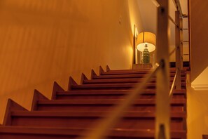 Uma escadaria com degraus de madeira e corrimão, dando acesso a um quarto com uma luminária.
