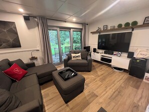 A modern living room with a grey sofa, a grey armchair, and a black ottoman. There is a flat-screen TV mounted on the wall, a white entertainment unit, and a large window with curtains. - Country House for Winter Sports (Magog)