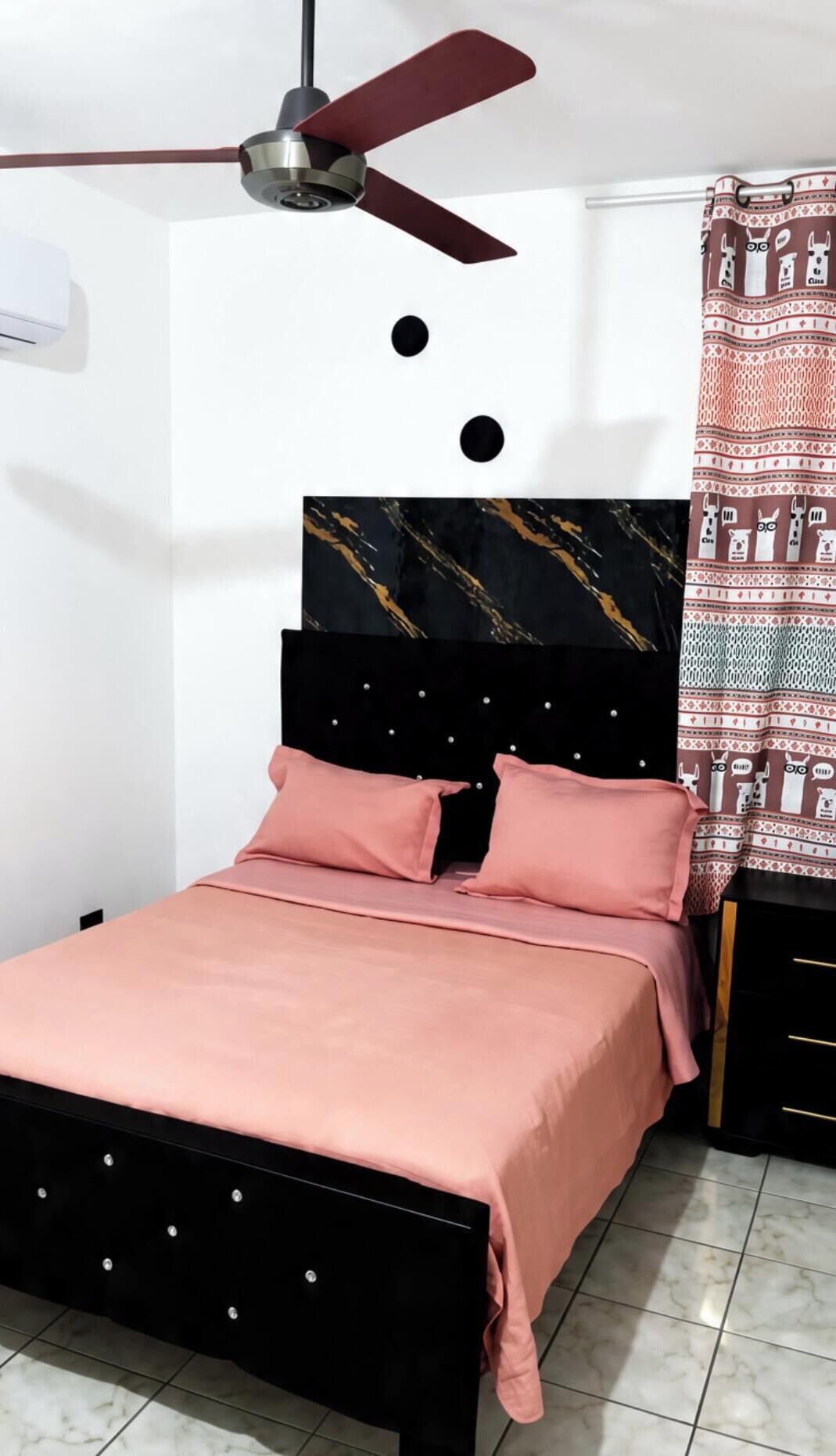 A bedroom with a bed, a ceiling fan, a nightstand, and a patterned curtain.