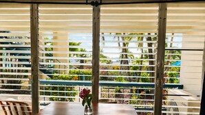 Un comedor con una mesa y sillas, un jarrón con flores y vistas a través de una ventana con persianas horizontales.